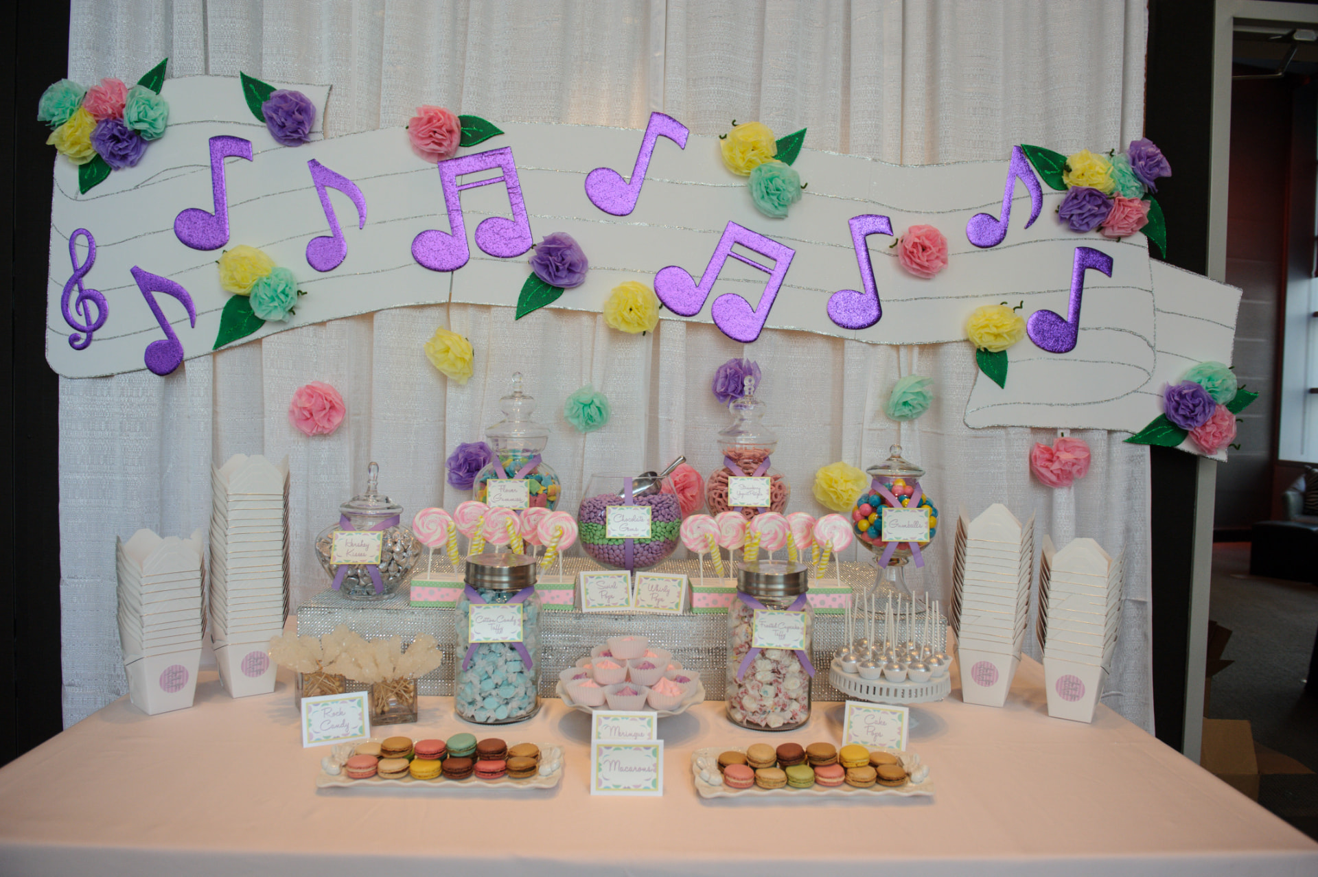 A first birthday dessert table with purple musical notes, pastel paper flowers and jars of sweets