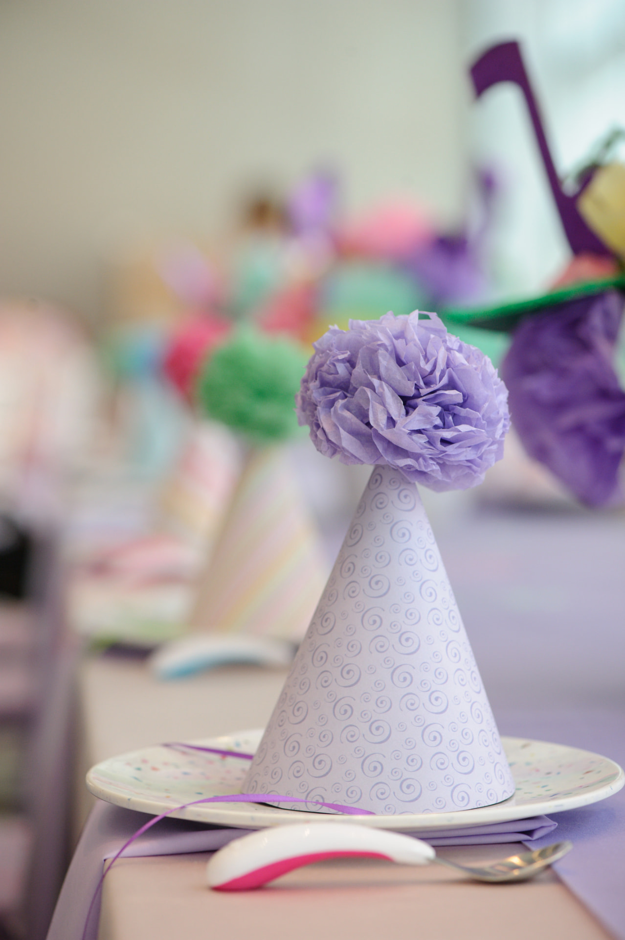 A lavender party hat topped with a tissue-paper flower at a birthday place setting